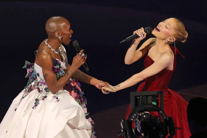 Two performers on stage; one in a floral-accented gown, the other in an elegant strapless dress. Both holding microphones and singing passionately