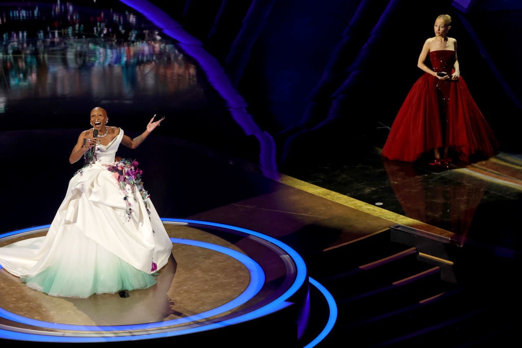 Two people on stage at an event. The person on the left is singing in a gown with floral details. The person on the right wears a dark red gown