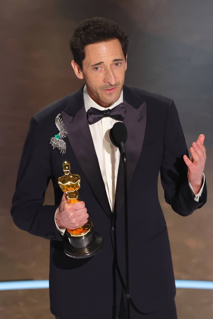 Adrien Brody in a formal suit holding an Oscar statue and speaking on stage
