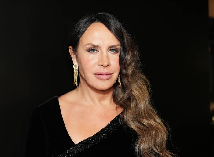 Karla Sofia Gascon with long wavy hair, wearing an elegant black dress with a V-neck and long earrings, poses against a dark background
