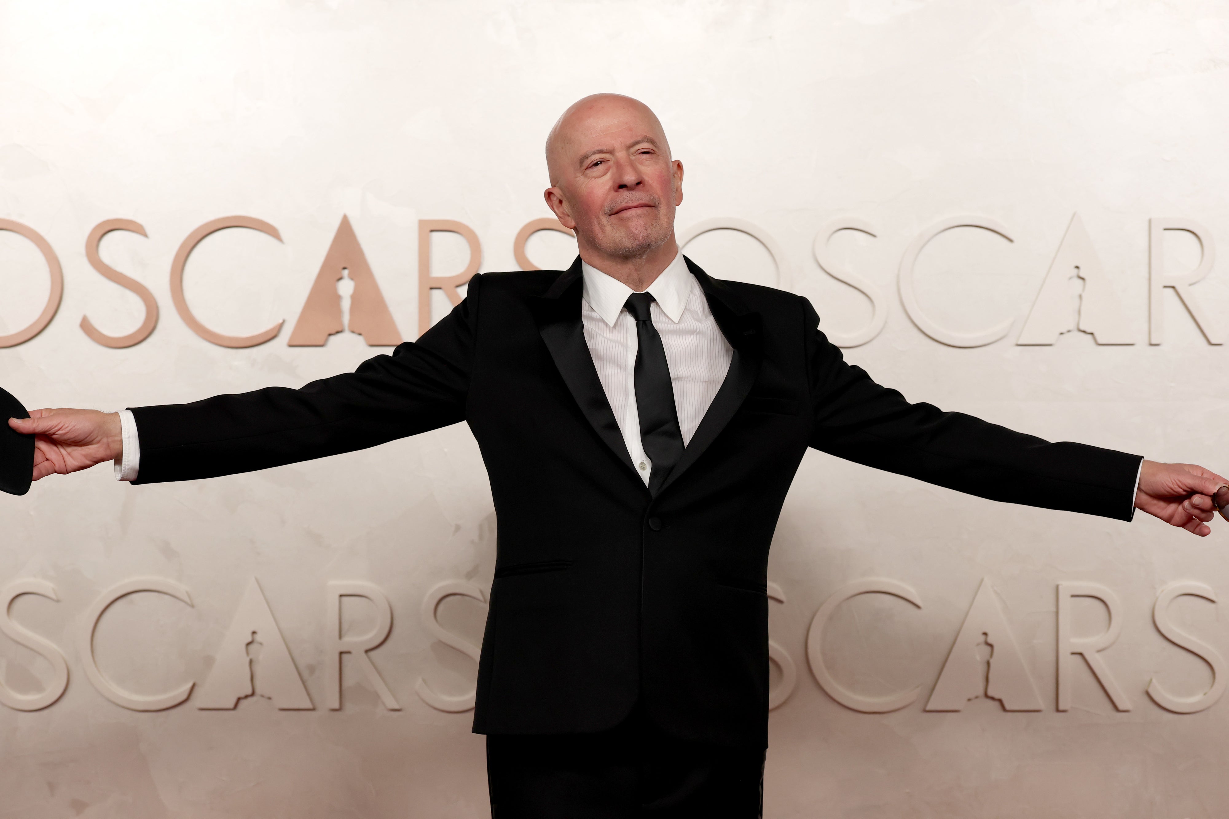 Jacques in a black tuxedo posing with arms outstretched at an event. The backdrop features large text reading "OSCARS."