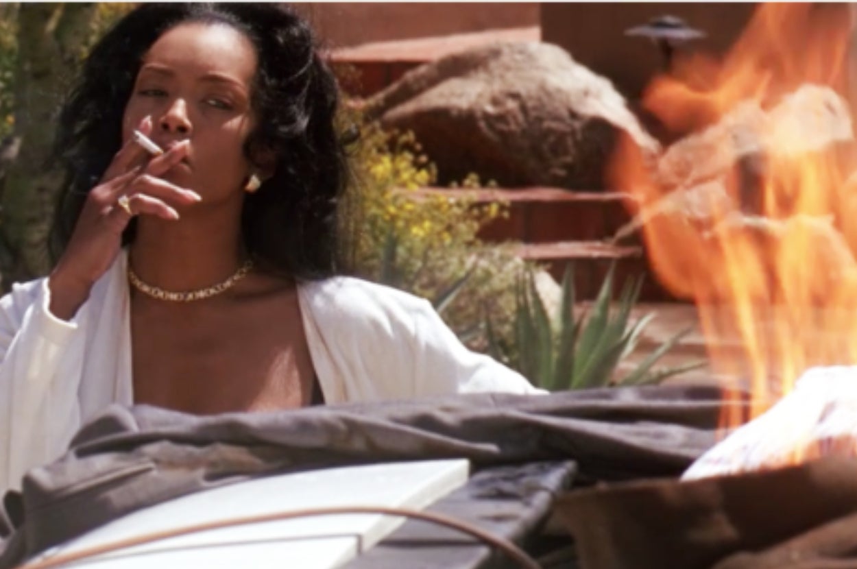 Person with long hair smokes a cigarette near an outdoor fire, dressed in a white robe