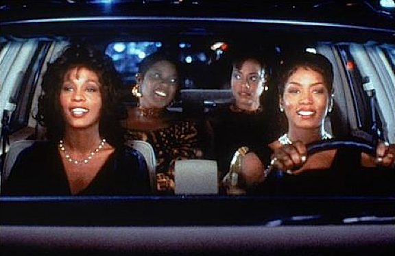 Four women in elegant attire are inside a car, appearing joyful and engaged in conversation