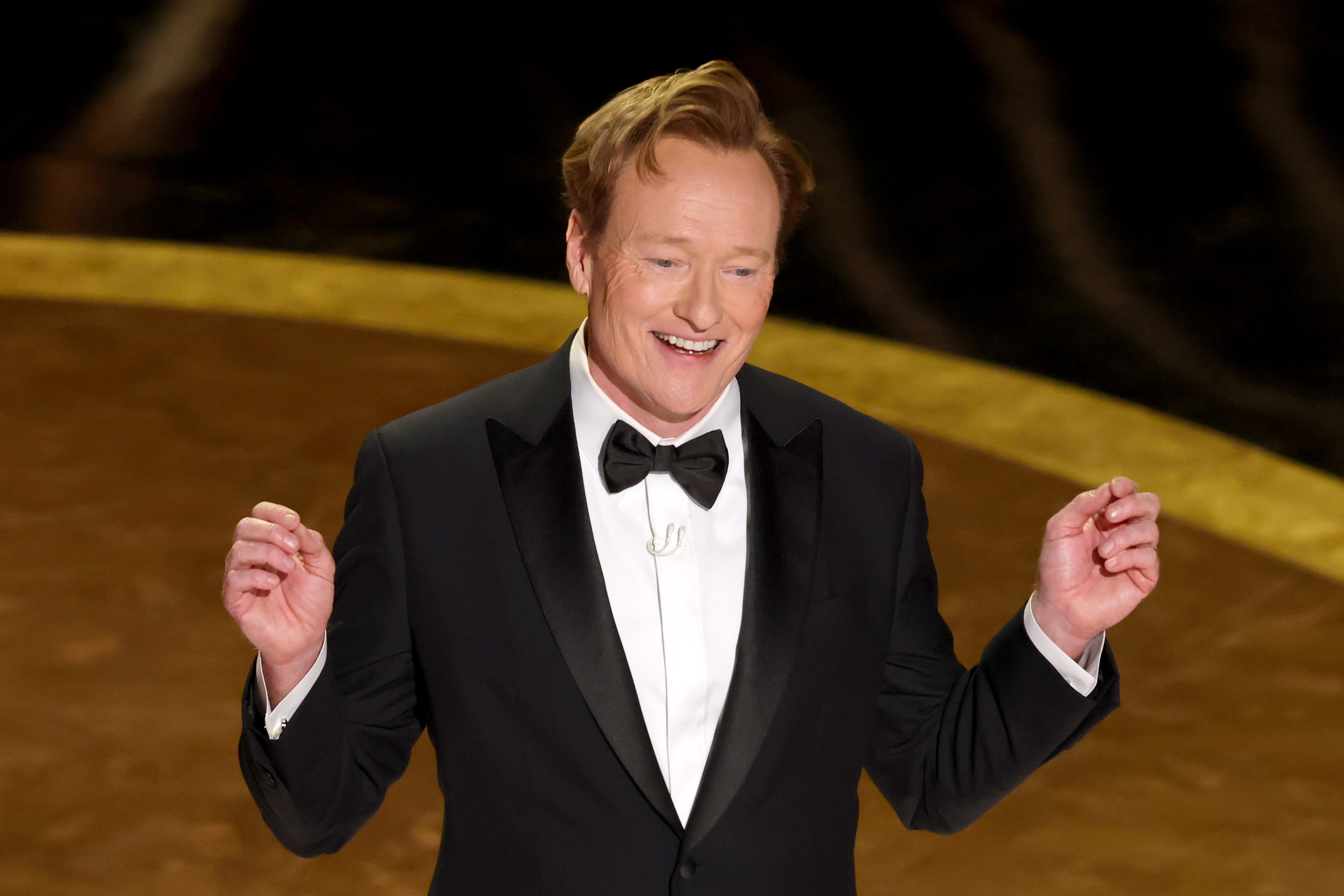 Person in a tuxedo on stage, smiling and gesturing with both hands
