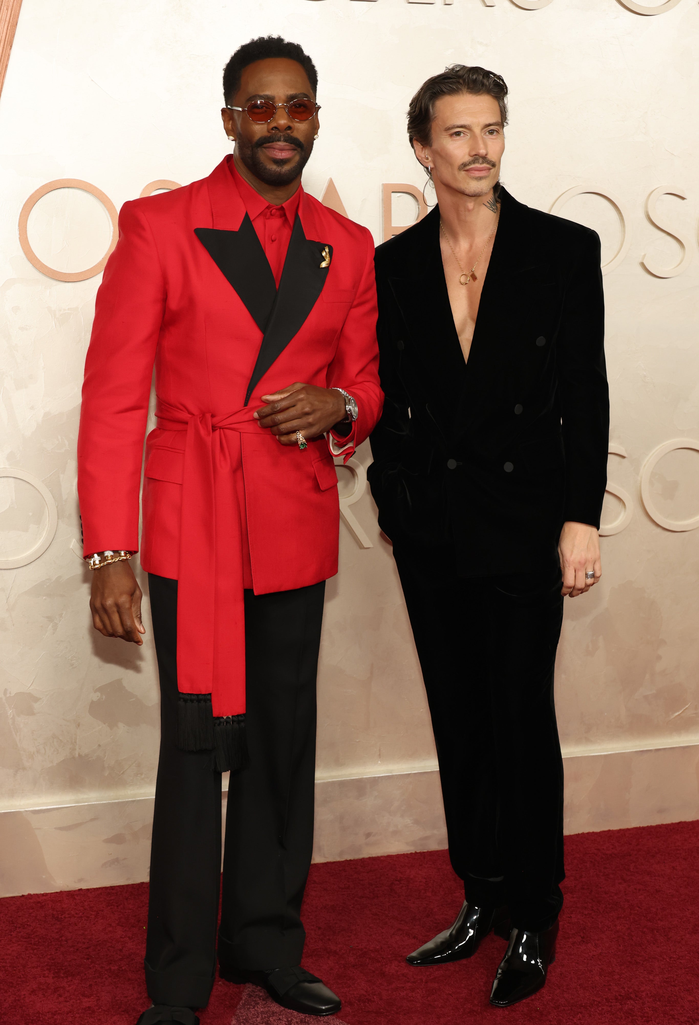 Colman Domingo and husband Raúl pose on a red carpet. One wears a tailored red suit, the other a velvet black suit with an open shirt