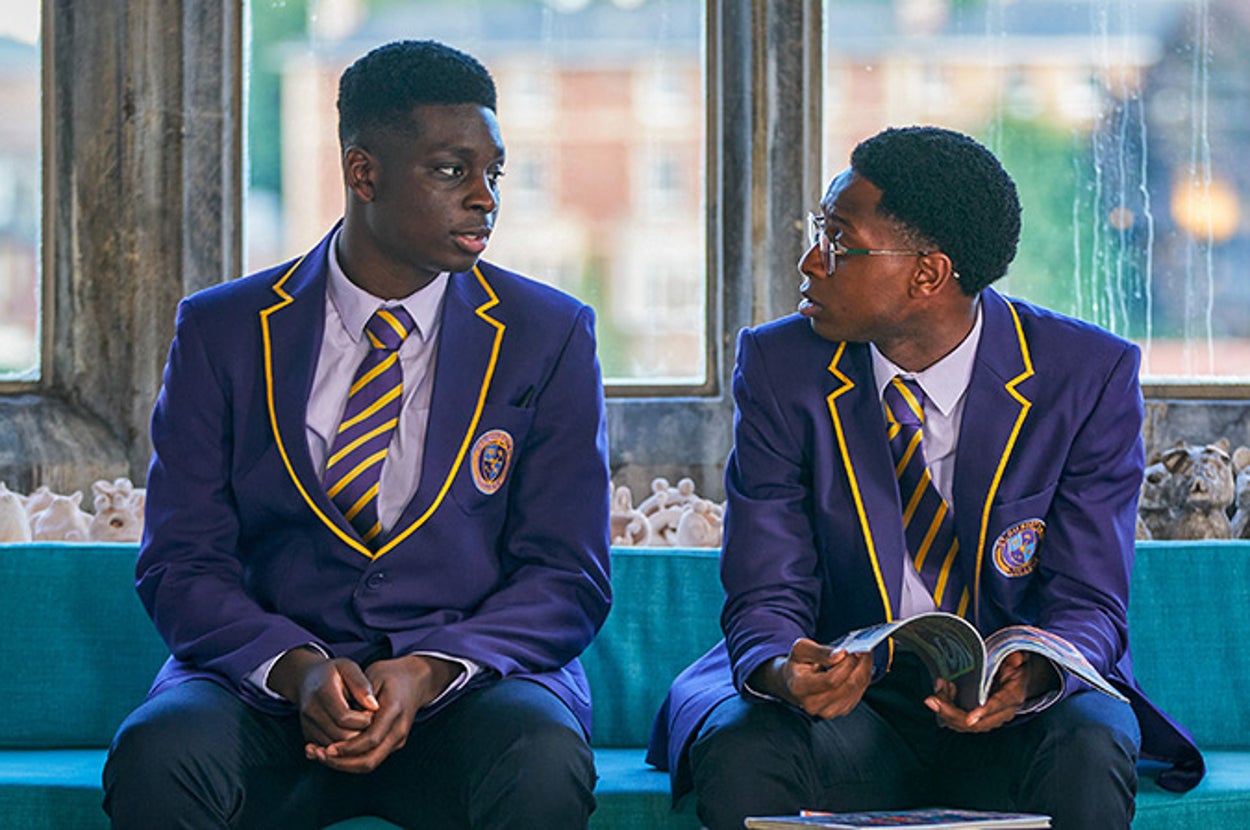 Two students in school uniforms sit on a bench talking. One holds a book, suggesting a school setting or discussion