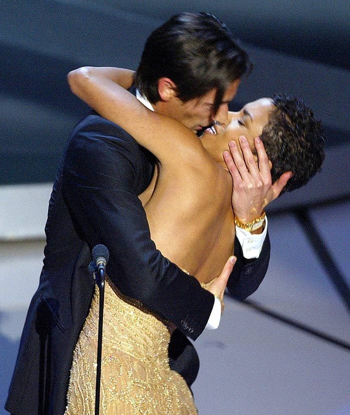 Two people embrace on stage, one in a backless gown and the other in a suit, appearing to celebrate at an event