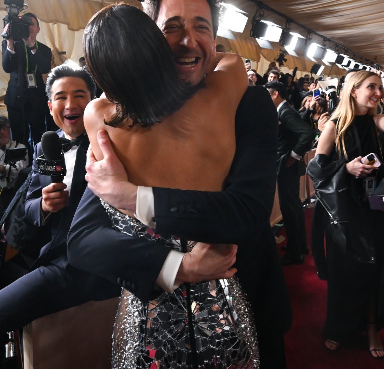 People embrace joyfully on the red carpet, wearing elegant formal attire, surrounded by photographers capturing the moment