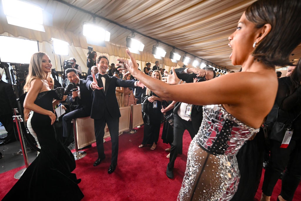 Celebrities greet each other on a red carpet. They wear elegant, form-fitting gowns and tuxedos in an event setting with photographers in the background