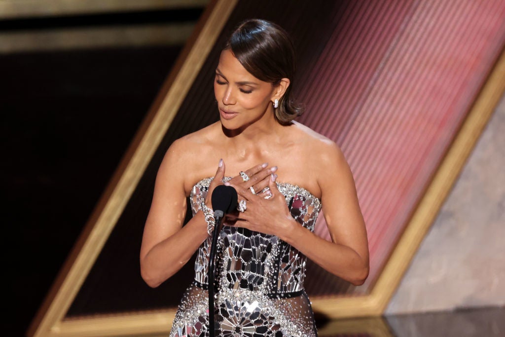 A woman stands at a microphone wearing a strapless, sequin-covered dress, hands on her chest, possibly making a speech at an event