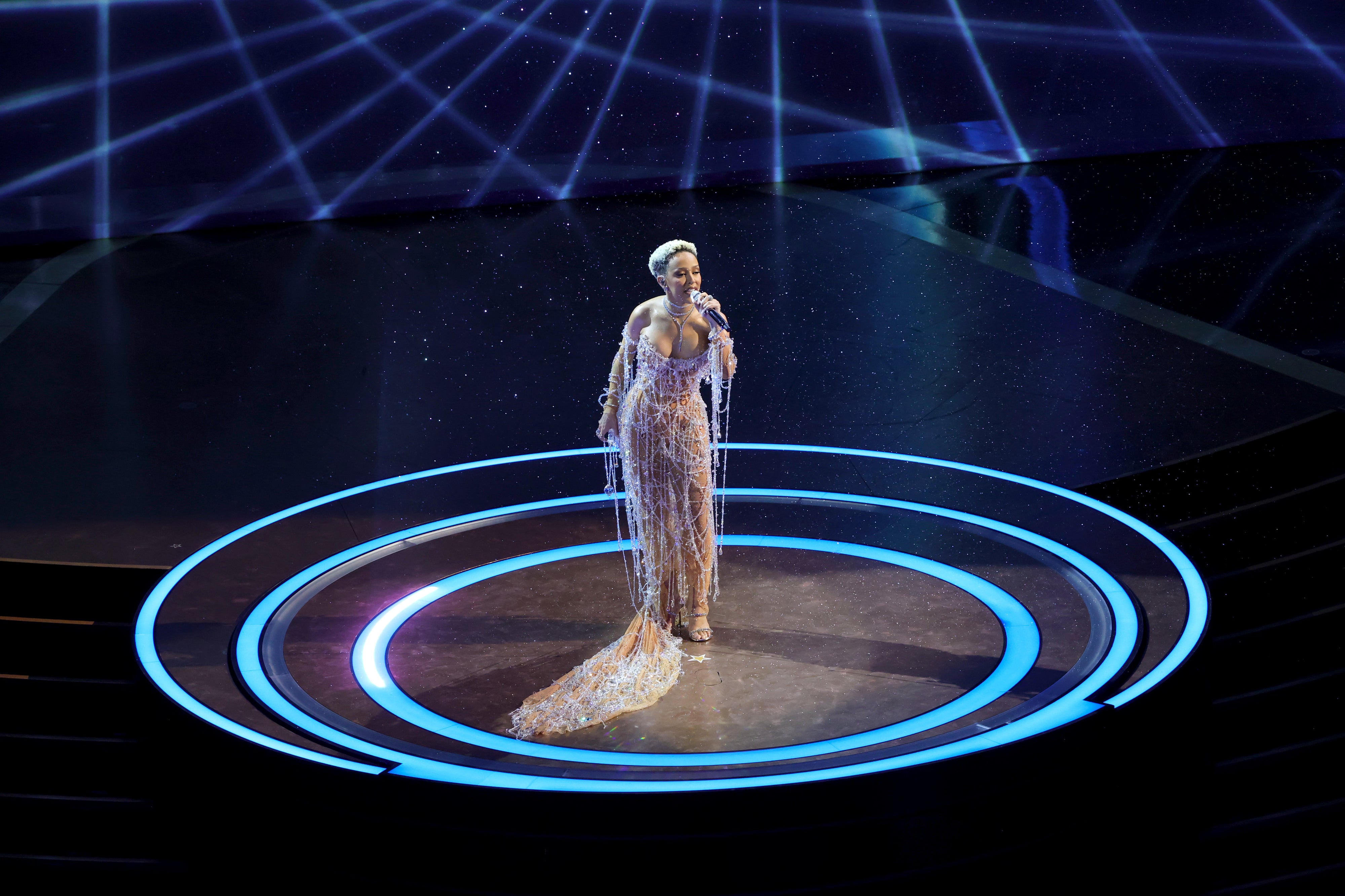Singer performing on stage in an elegant, sparkling gown with geometric light patterns in the background