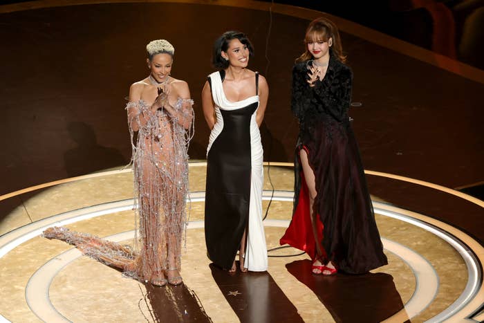 Three celebrities in glamorous gowns stand together on stage, holding microphones, during a live event