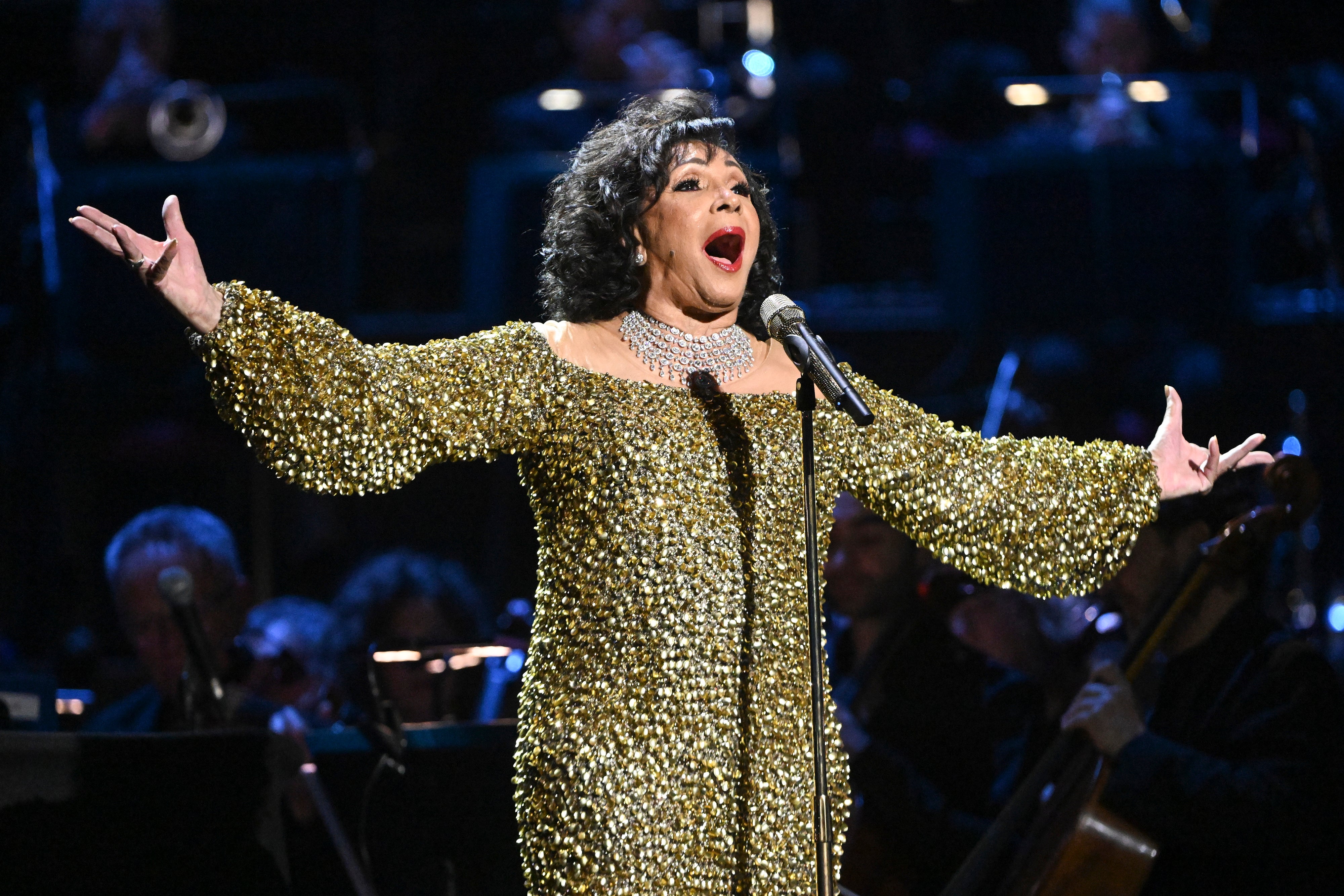 A performer in a sparkling gown sings passionately on stage with an orchestra in the background