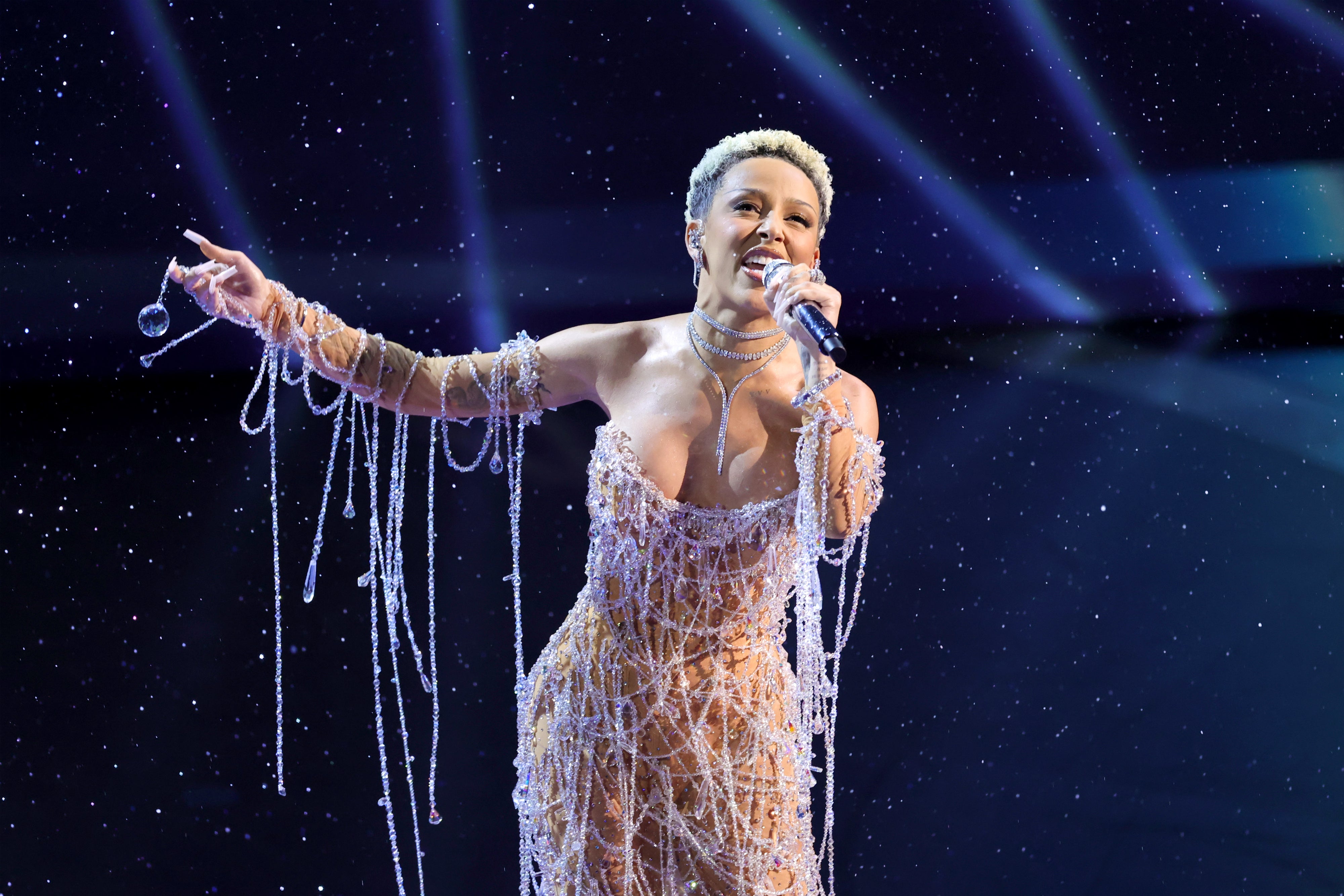 A performer on stage wearing an ornate, crystal-adorned gown, holding a microphone and gesturing with their other hand amidst a starry backdrop