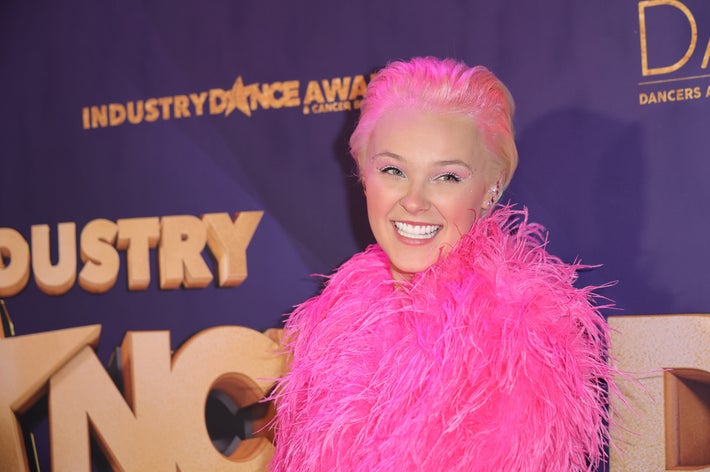 JoJo Siwa smiling at an event, wearing a large feathered pink jacket