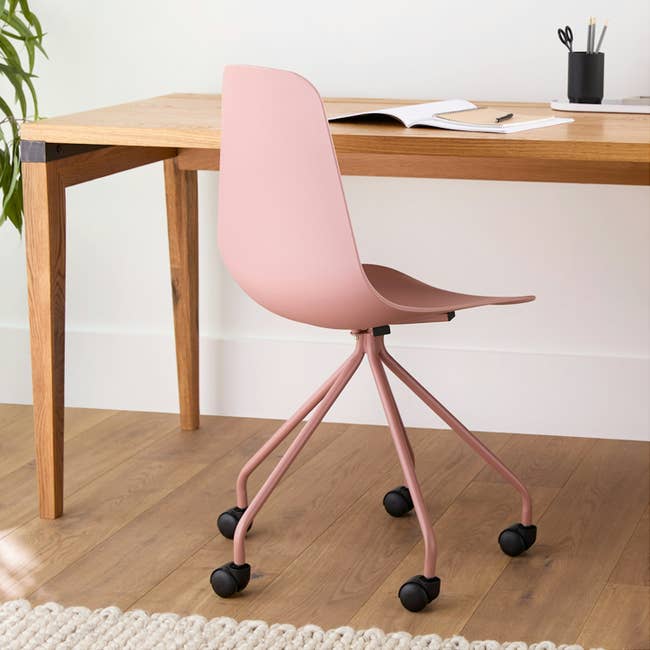 A modern workspace with a pink chair on wheels, a wooden desk, and notebooks and pens arranged neatly on top