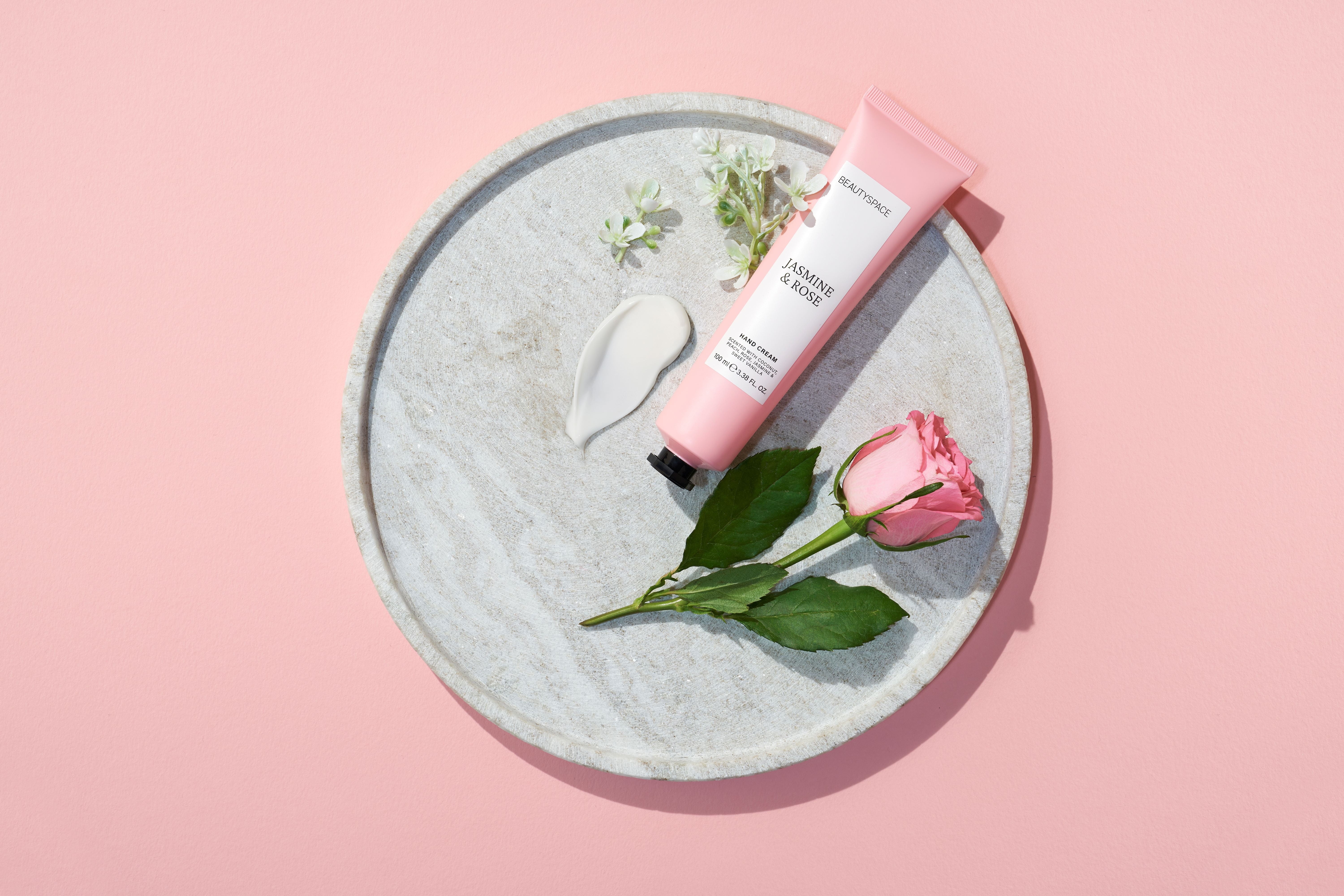 The pink tube of hand cream with a black twist top next to a rose and a dollop of cream showing its smooth texture