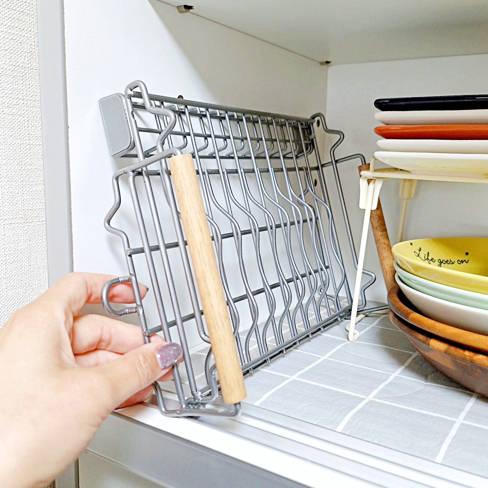 手を使ってキッチンの棚に金属製の整理ラックを置く様子。隣には皿が積み重なっている。