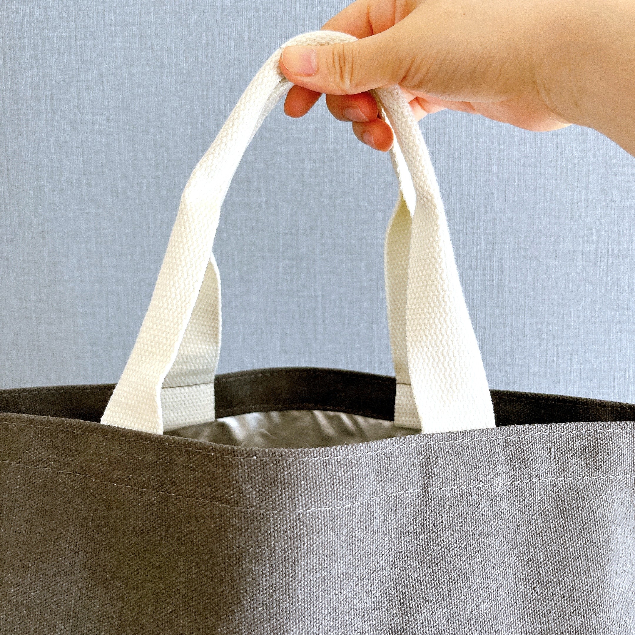 白い持ち手のトートバッグを指で持ち上げている様子。バッグの素材感が伝わるシンプルなデザイン。