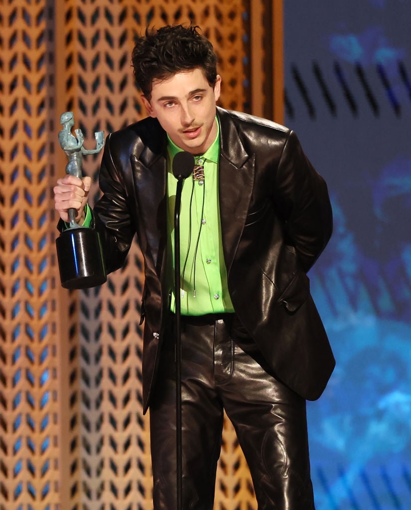 Person in a shiny leather suit with a bright shirt holds a trophy while speaking into a microphone at an award event