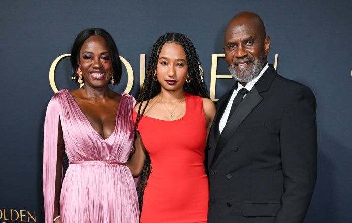 Three people pose on the red carpet at the Golden Globes. Left person in a pleated gown, middle in a sleek dress, right in a classic tuxedo
