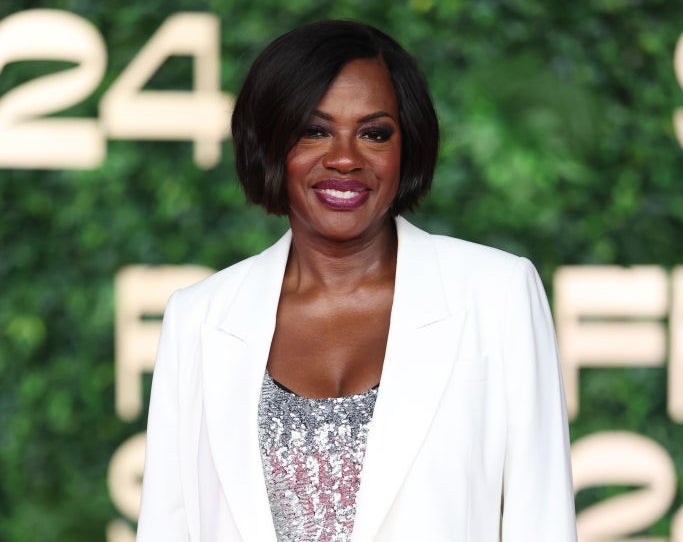 Celebrity in a white suit with a sequined top stands on a red carpet. A green backdrop with large text is visible behind them