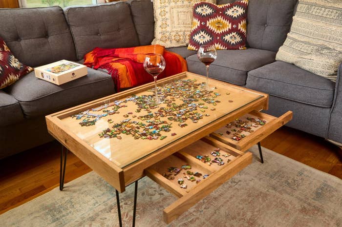 Coffee table with a puzzle in progress, two wine glasses, and pull-out puzzle trays, placed between a comfy couch and colorful pillows