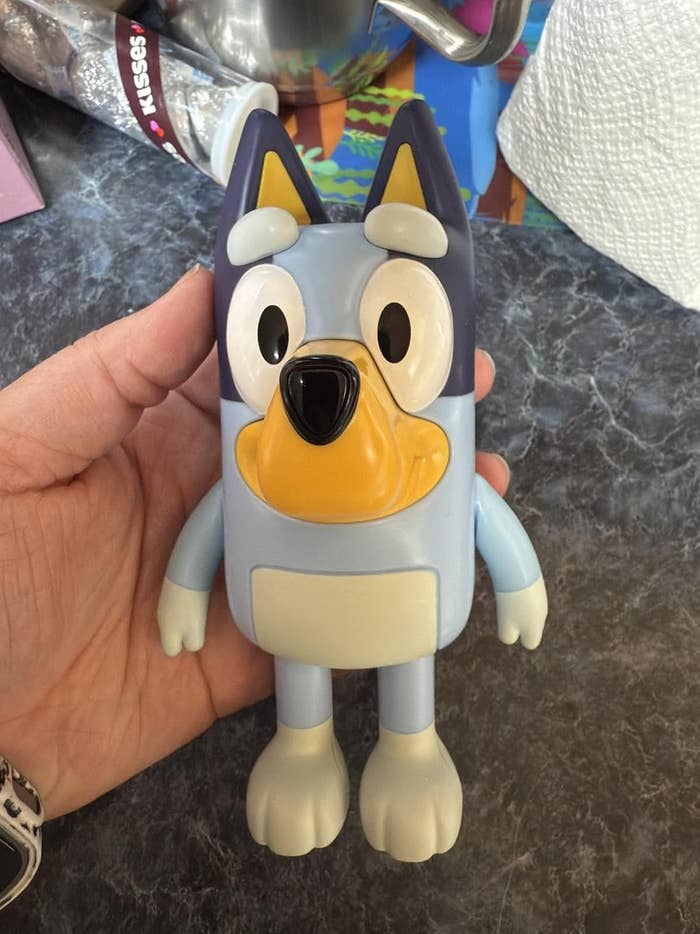 Hand holding a cartoon quality artifact resembling an animated bluish dog, with floppy ears and expressive eyes, suitable for children's merchandise