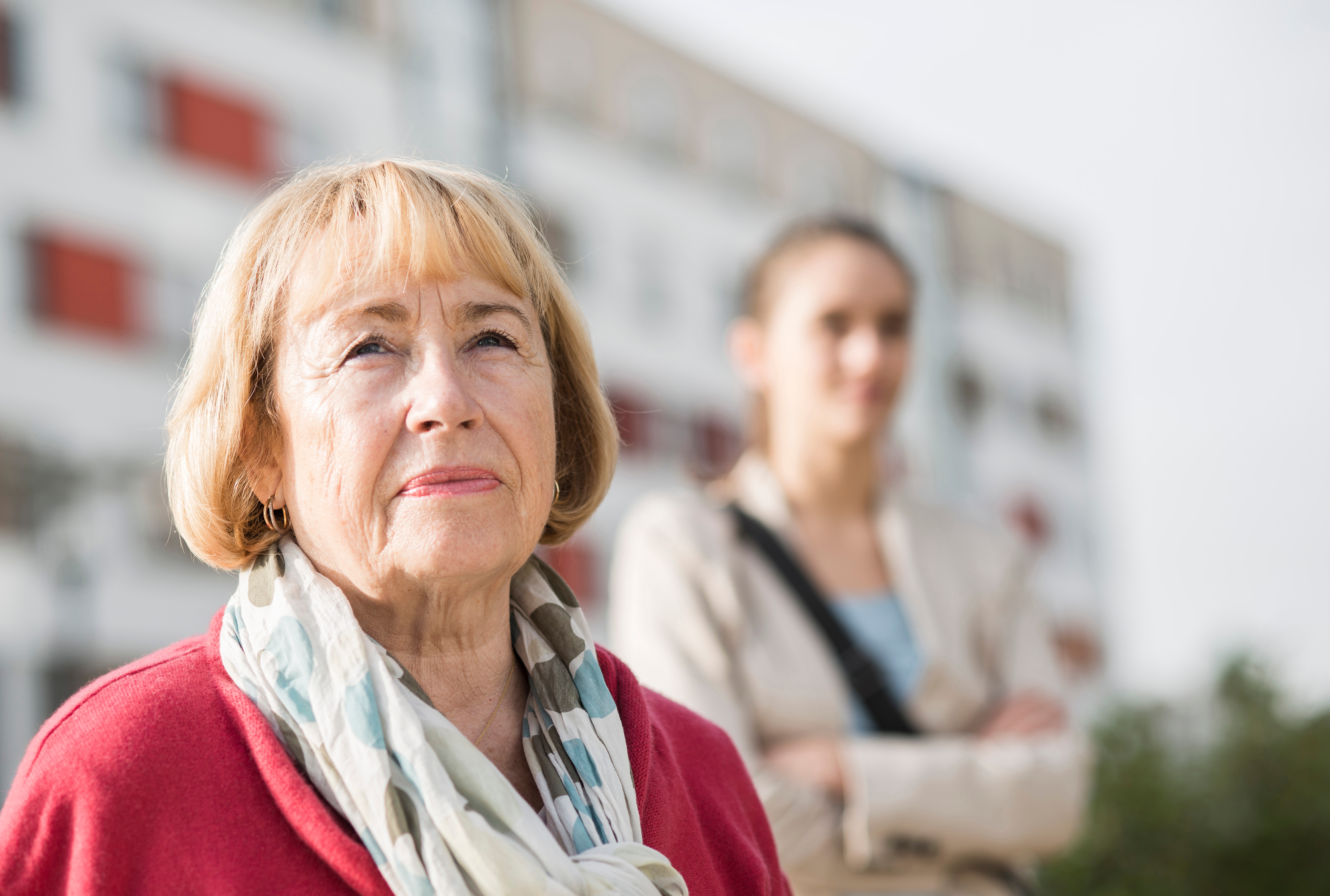 En eldre kvinne ser gjennomtenkt inn i det fjerne, iført et skjerf og en genser. En uskarp person står i bakgrunnen