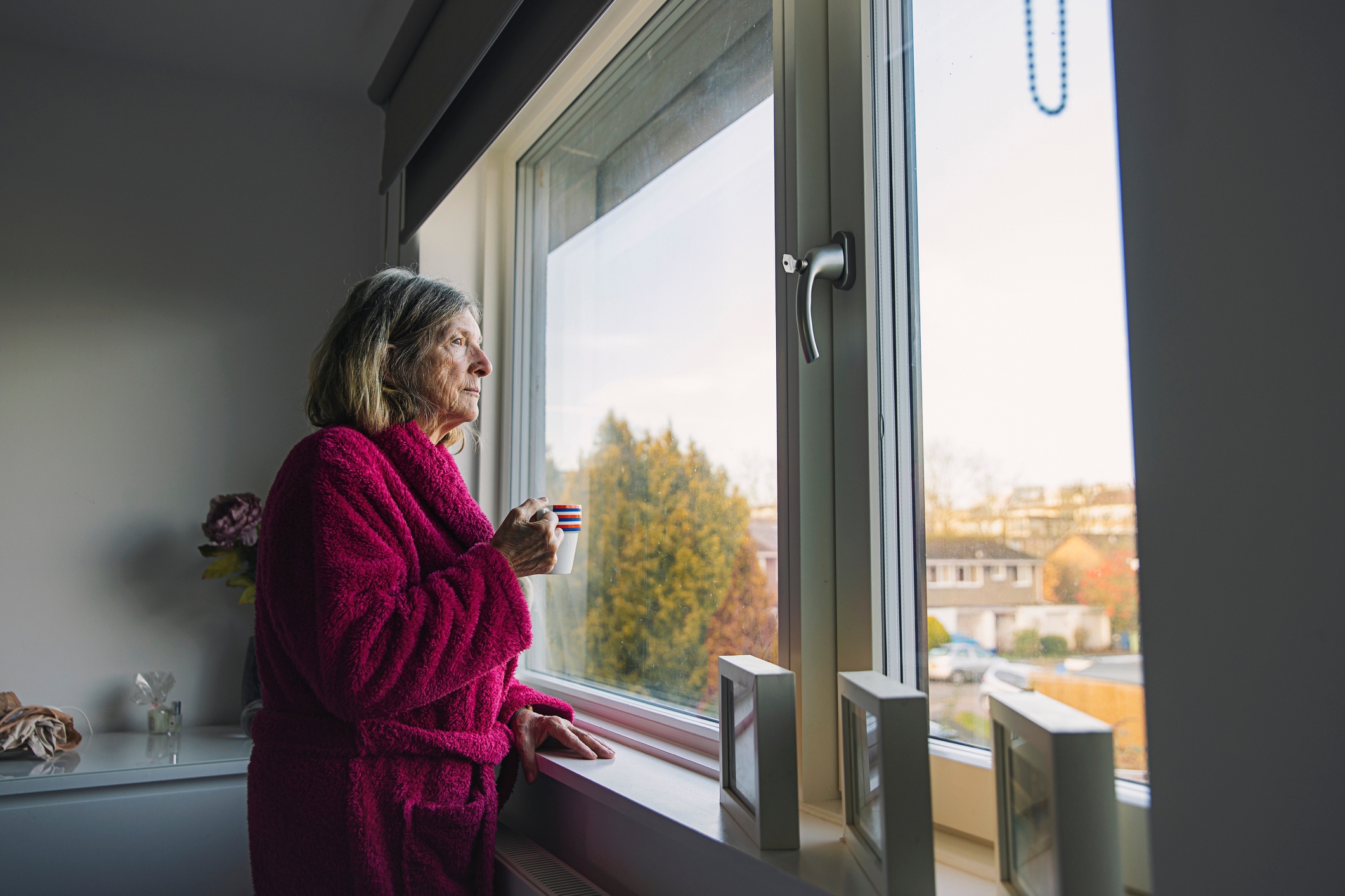 Eldre kvinne i en koselig kappe ser ut av et vindu mens han holder et krus, og fremstår som fredelig og kontemplativ