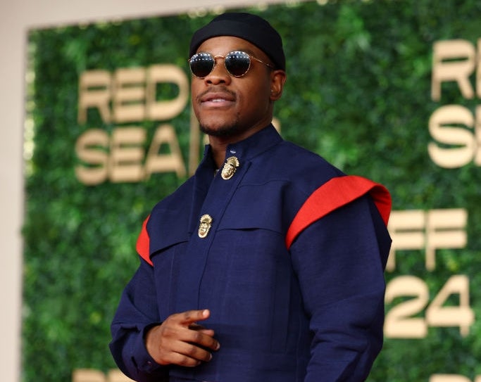 Person on red carpet in navy jacket with red accents, wearing sunglasses and round hat, standing in front of a green backdrop with "Red Sea" text