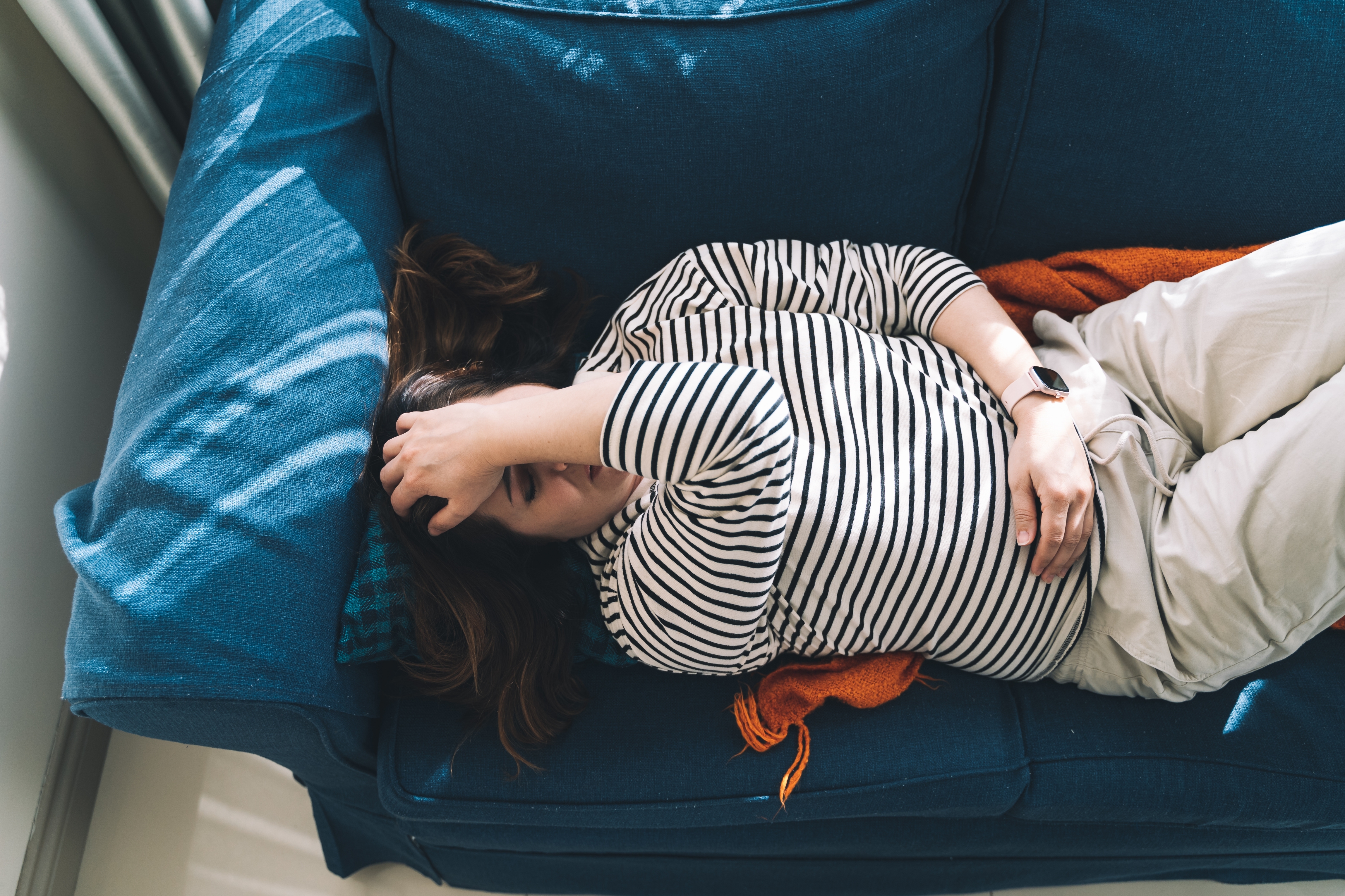 Person i stripete skjorte ligger på en sofa med en hånd på pannen og en annen på magen, og ser ut til å hvile eller lur