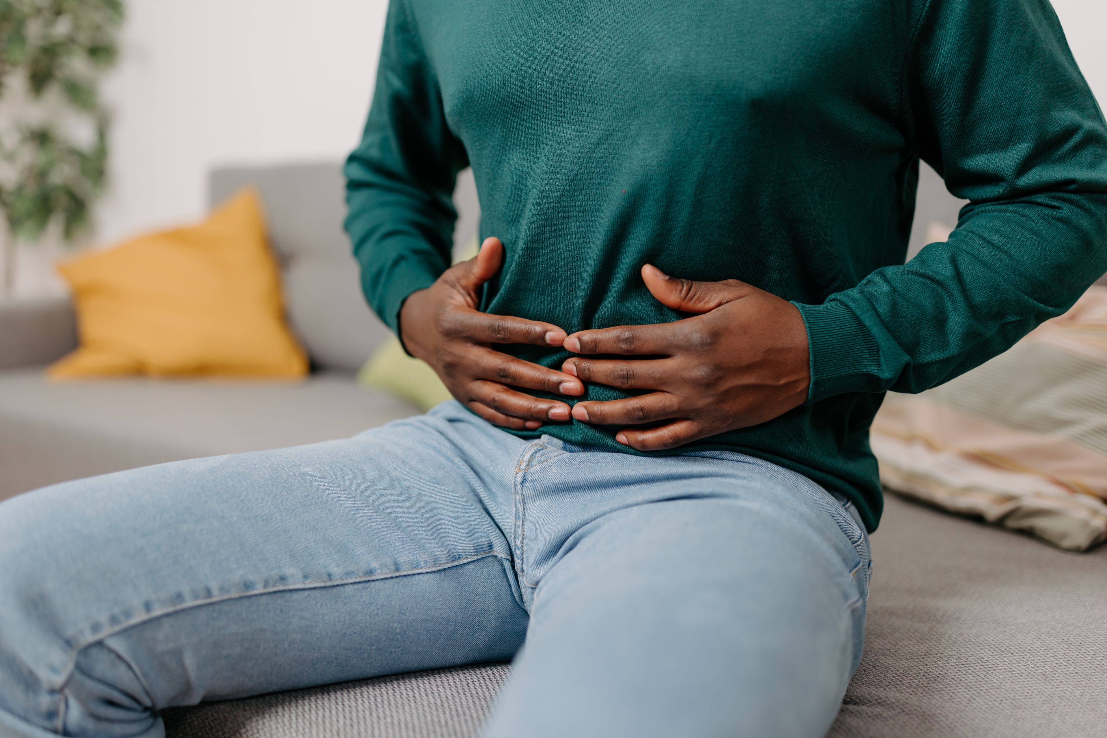 Person som sitter på en sofa som holder magen med begge hender, har på seg en skjorte og jeans med lang ermet og jeans