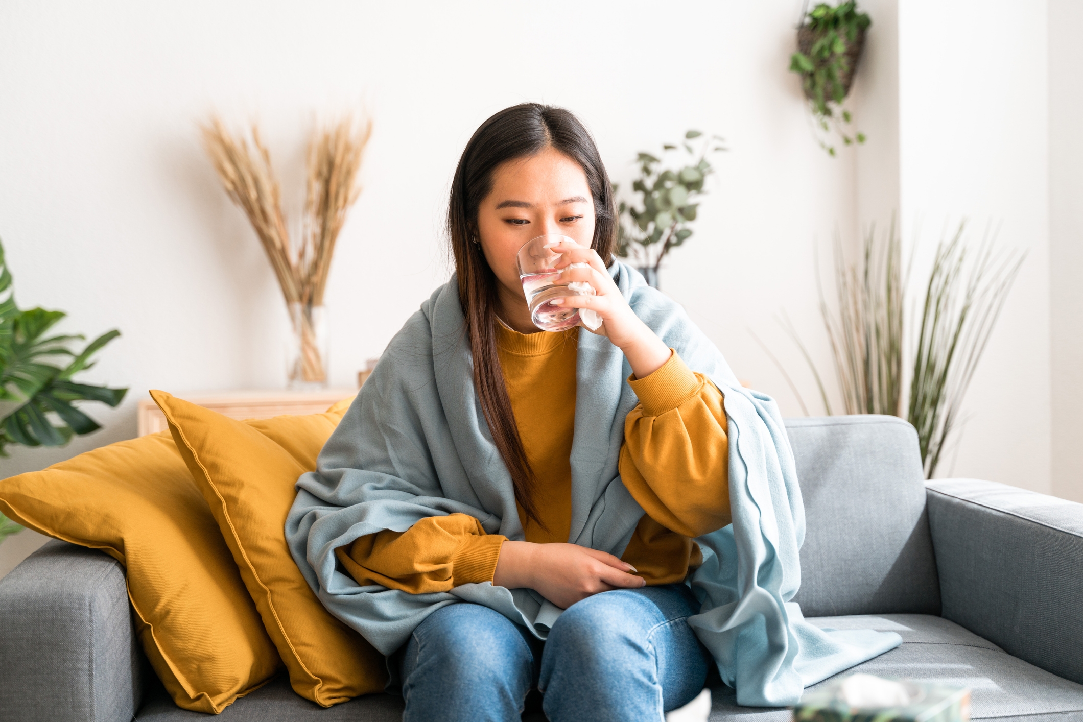 Person som sitter på en sofa, pakket inn i et teppe, drikker et glass vann, med planter og puter i bakgrunnen