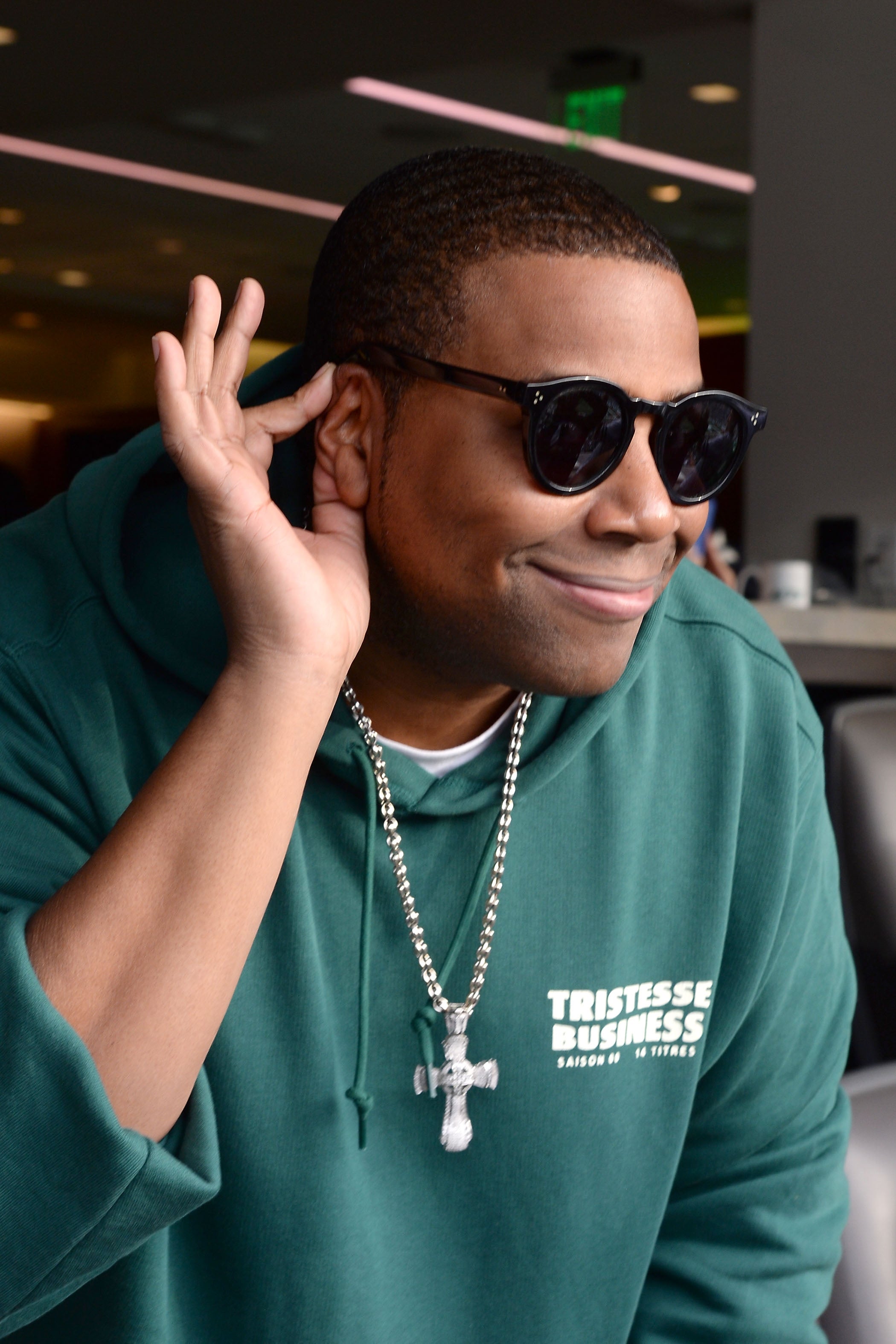 Kenan Thompson in a hoodie and sunglasses playfully cups their ear as if listening closely