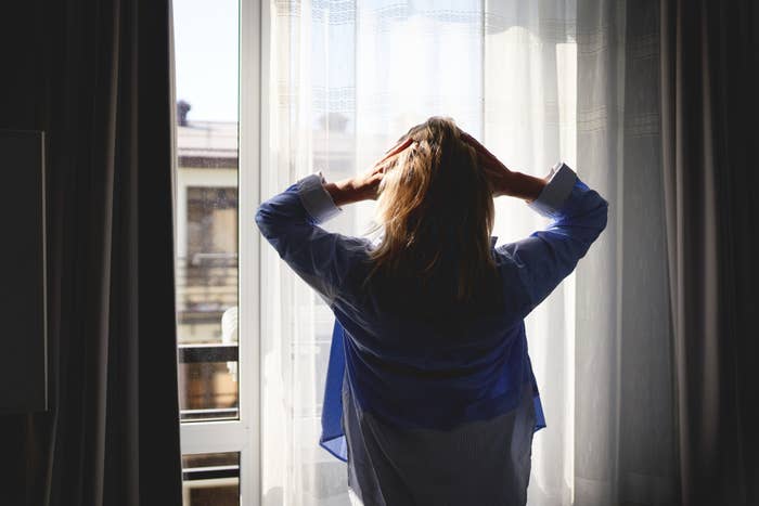 Person standing by a window, back to camera, holding their head, conveying contemplation or stress in a personal moment at home