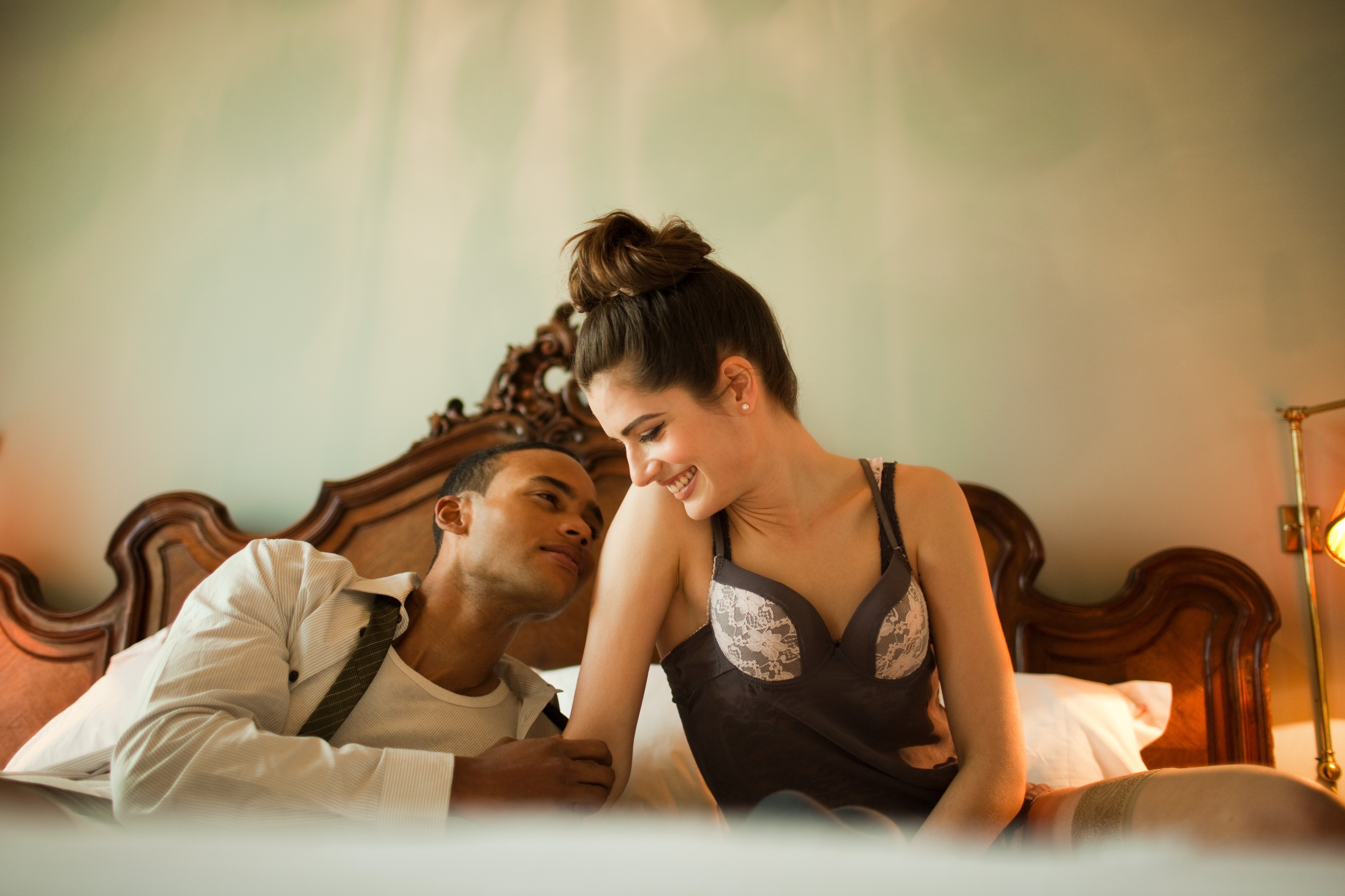 A couple sits on a bed, smiling at each other warmly. The woman wears a camisole; the man is in an open shirt, creating a relaxed, intimate setting