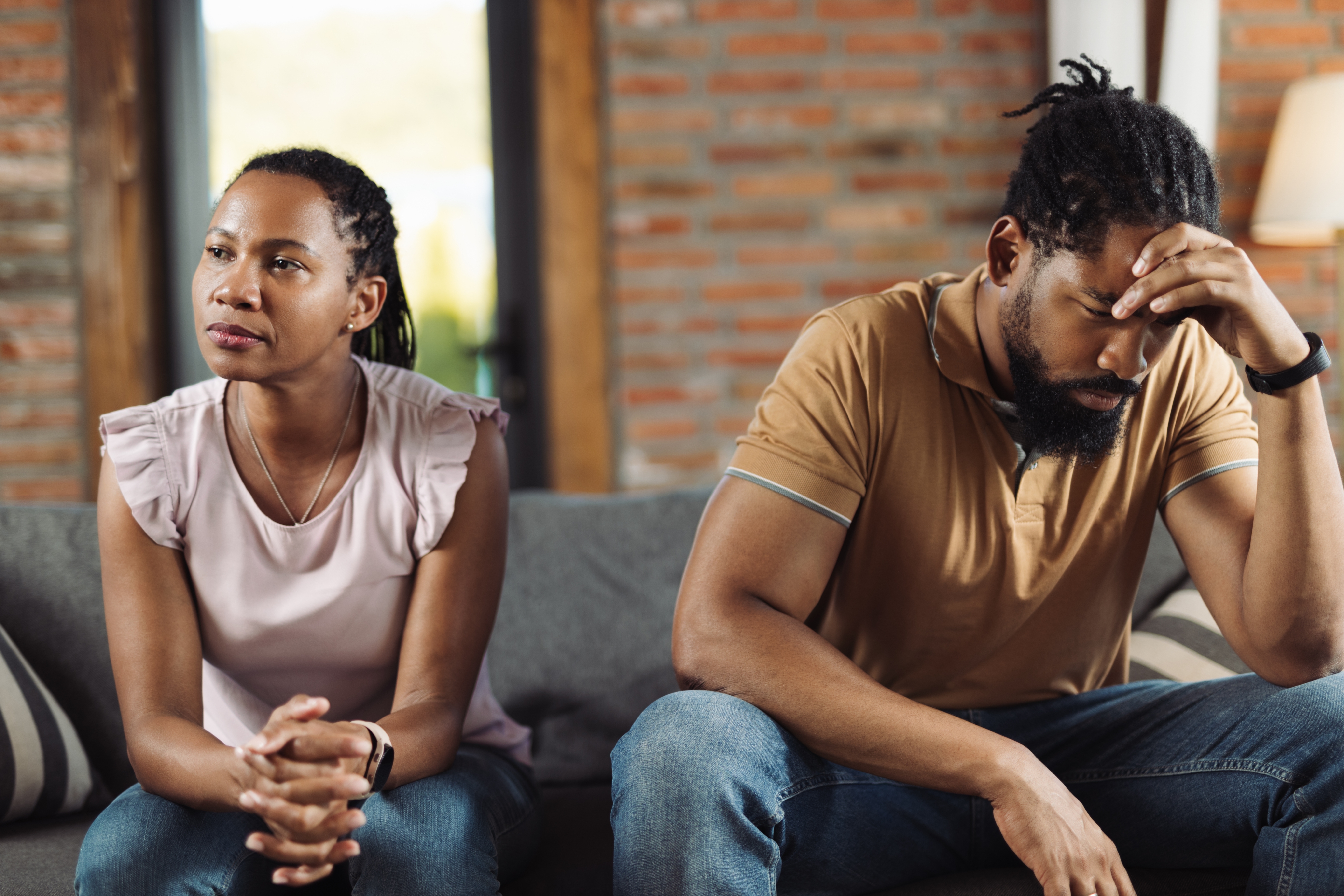 A couple sits on a couch in a tense moment; the woman looks away thoughtfully, while the man appears stressed, with his head in his hand
