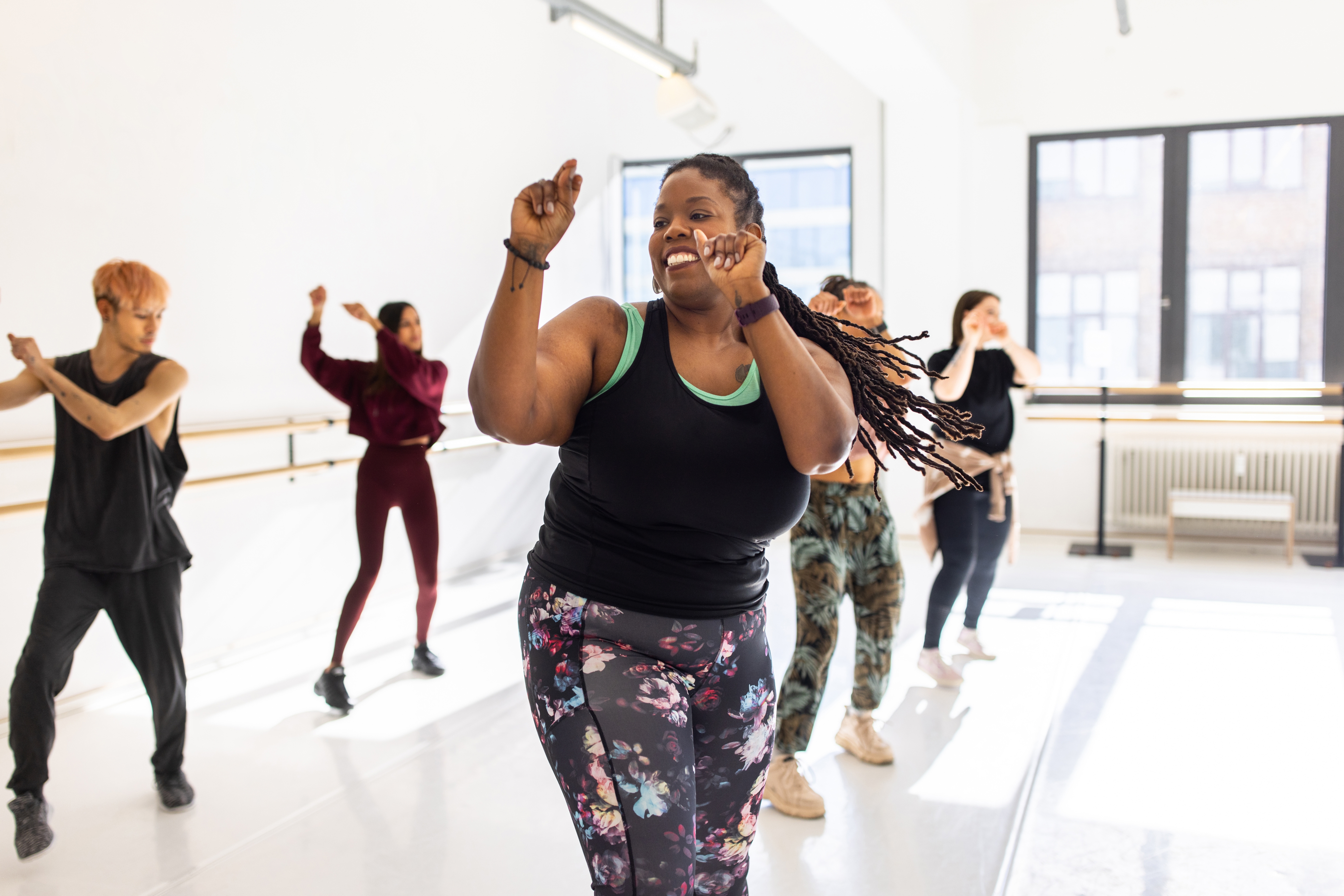 Folk danser gledelig i et studio, ledet av en smilende kvinne i en tank top og blomsterlegings