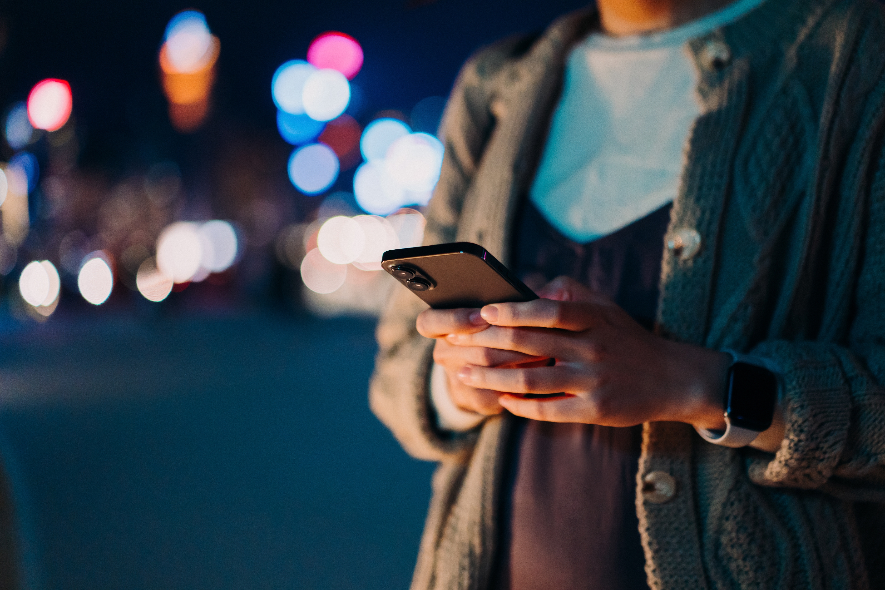 Person som sms på en smarttelefon i urbane omgivelser om natten, med uskarpe bylys i bakgrunnen