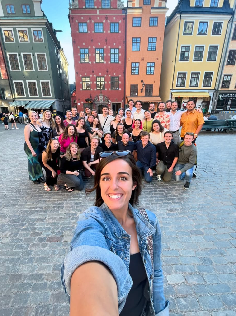 A group of people poses for a selfie in a lively city square with colorful buildings in the background