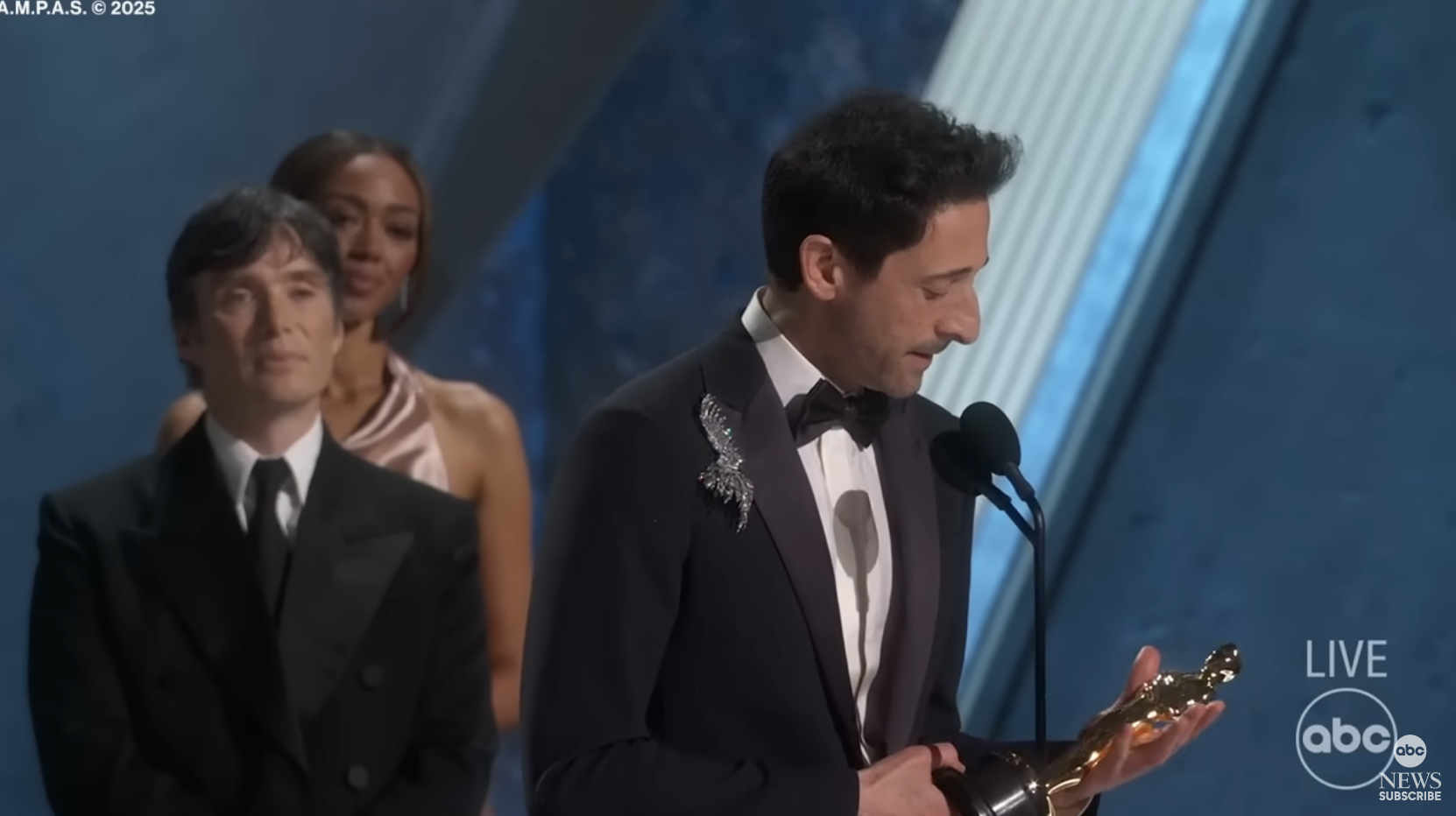 Person in formal wear holding an award on stage, with two others standing behind, also dressed formally, possibly during an awards ceremony