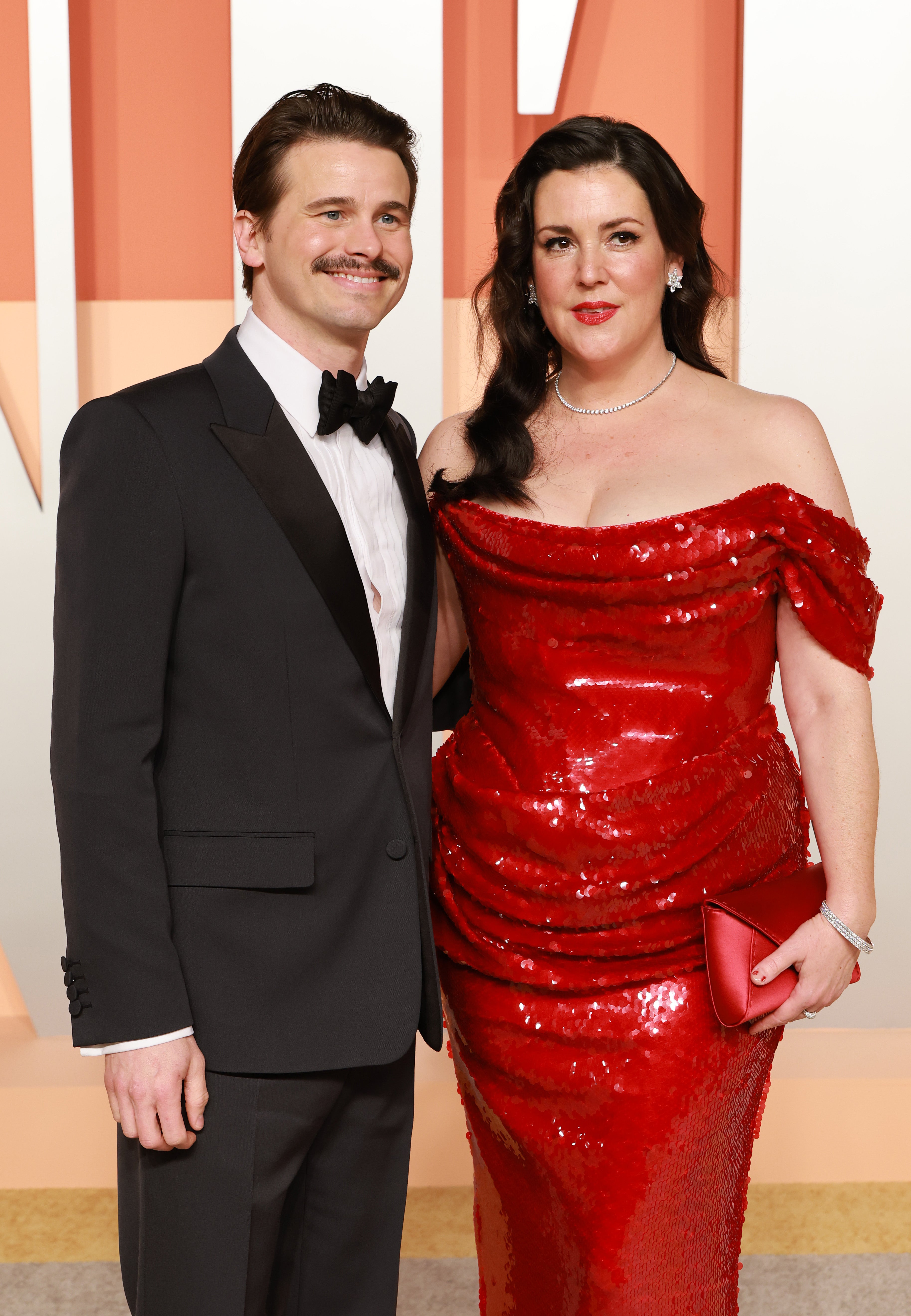 A man in a tuxedo and a woman in a red sequined off-shoulder gown pose together on a red carpet