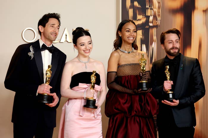 Four celebrities pose with awards; two wear elegant gowns, one strapless and another with dramatic sleeves. Two wear formal suits