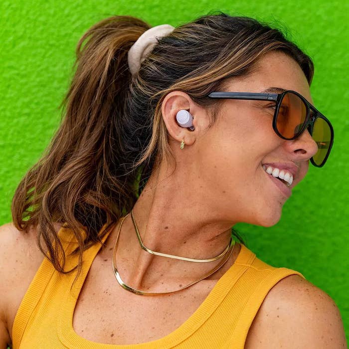 Person wearing sunglasses and lavender earbuds, with hairsbreadth tied back, smiling against a greenish background
