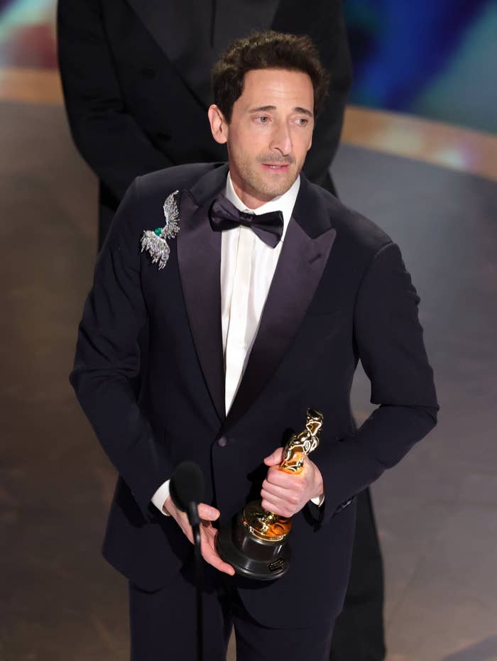 A person in a formal suit holding an award statue on a stage, appearing thoughtful, with a bird-shaped brooch on the lapel