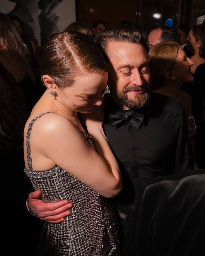 A couple embraces at a celebrity event, with the person on the left wearing an elegant, sleeveless dress with a textured pattern
