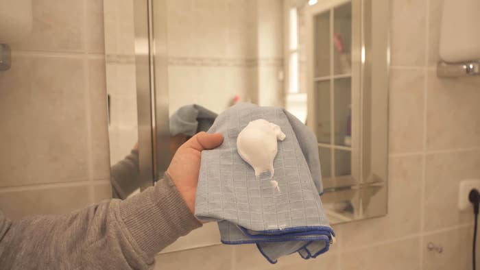 A person holds a cloth with a dollop of shaving cream in a bathroom, positioned in front of a mirror