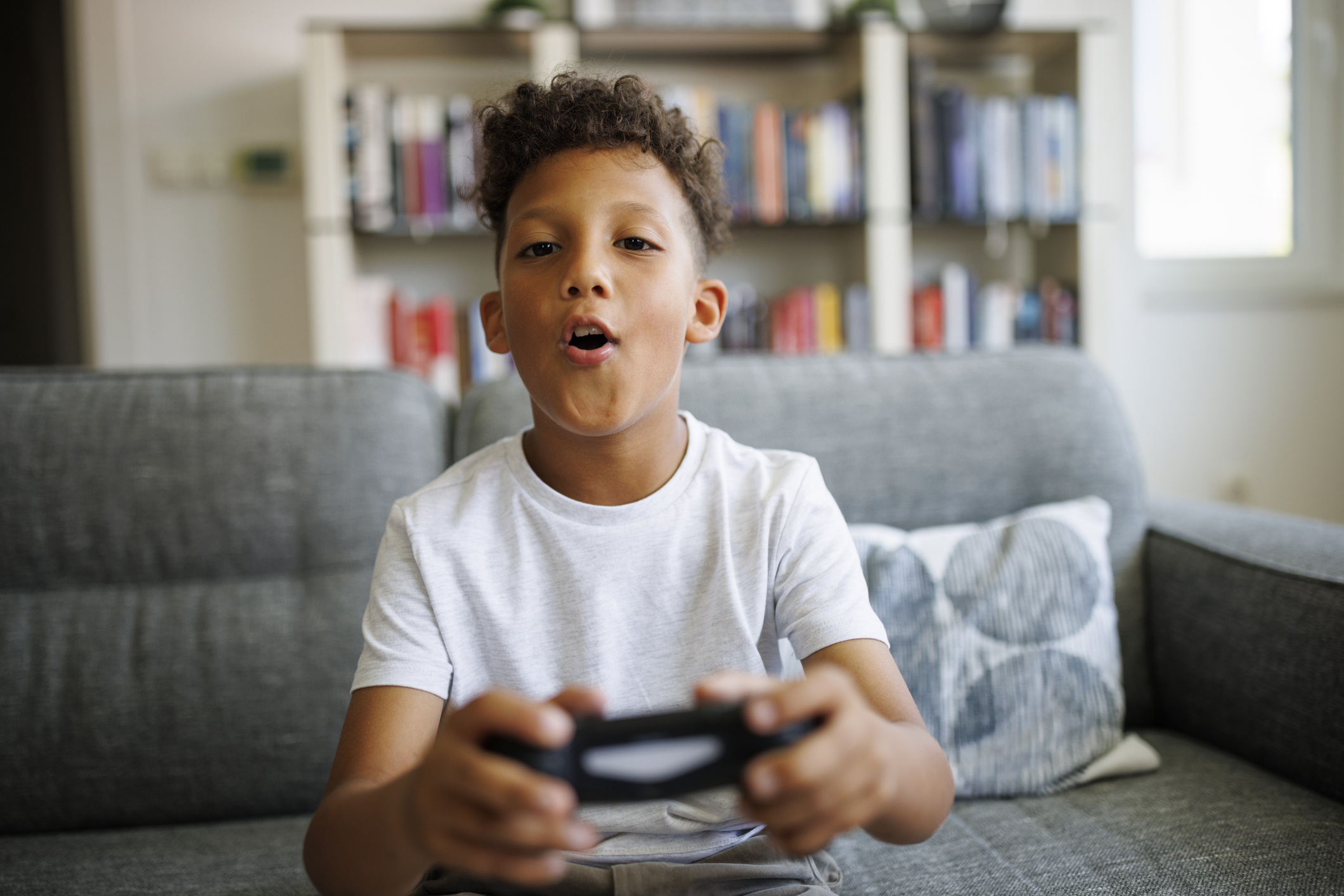 Barn som sitter på en sofa, med gledelig å spille videospill med en kontroller i hånden, hyller fylt med bøker i bakgrunnen