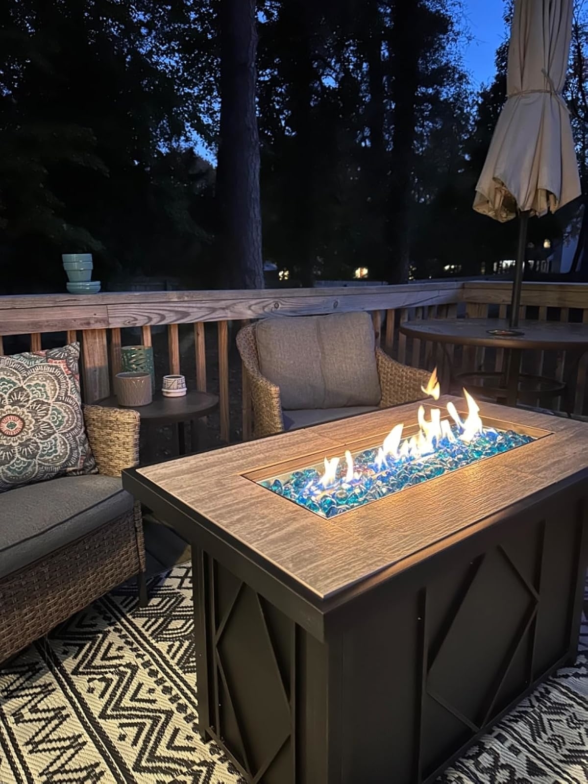 A cozy deck setup with a fire pit table, wicker chairs, patterned cushion, and an umbrella, perfect for outdoor relaxation or entertaining